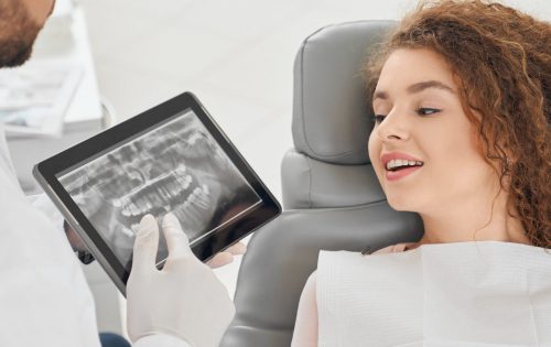 Front view of skilled male dentist in white uniform showing x ray picture of teeth to pretty woman. Female client sitting on chair and looking at tablet. Concept of treatment and health.